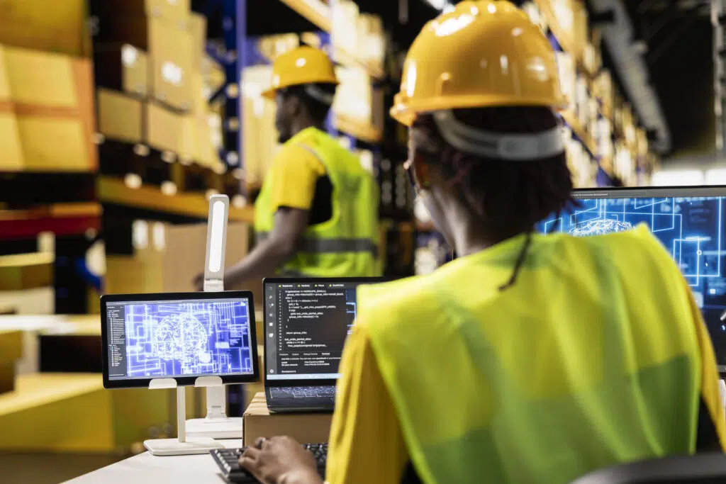 Funcionária de capacete e colete no computador vendo como funciona o sistema WMS em um armazém.