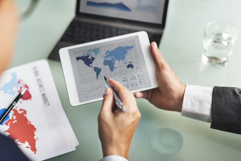 Uma mão segurando um tablet com um mapa mundial na tela e uma outra mão apontando uma caneta para um ponto do mapa, representando a logística de importação.