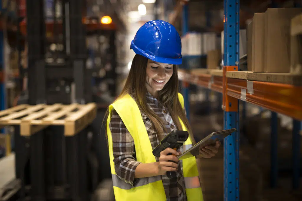 Mulher usando capacete azul e colete de segurança amarelo em um armazém, segurando um coletor de dados e uma prancheta, realizando controle de estoque com tecnologia RFID.
